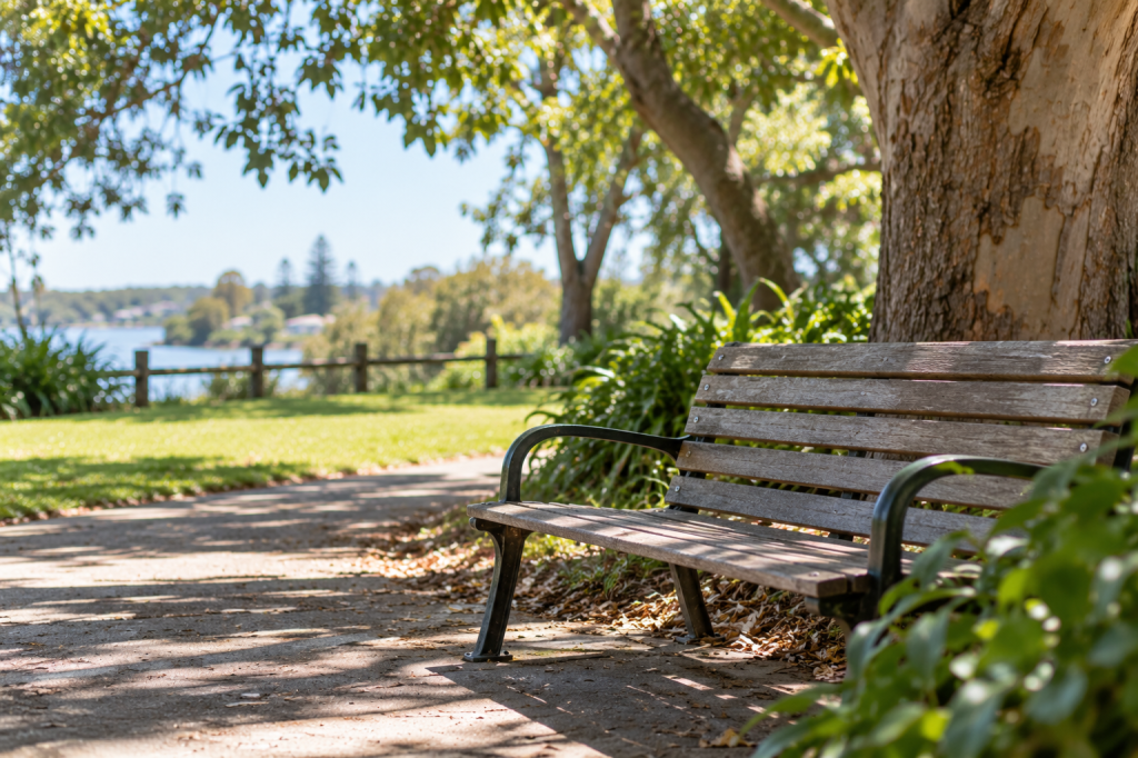 Empty wooden park bench beneath a leafy tree overlooking a peaceful waterfront.